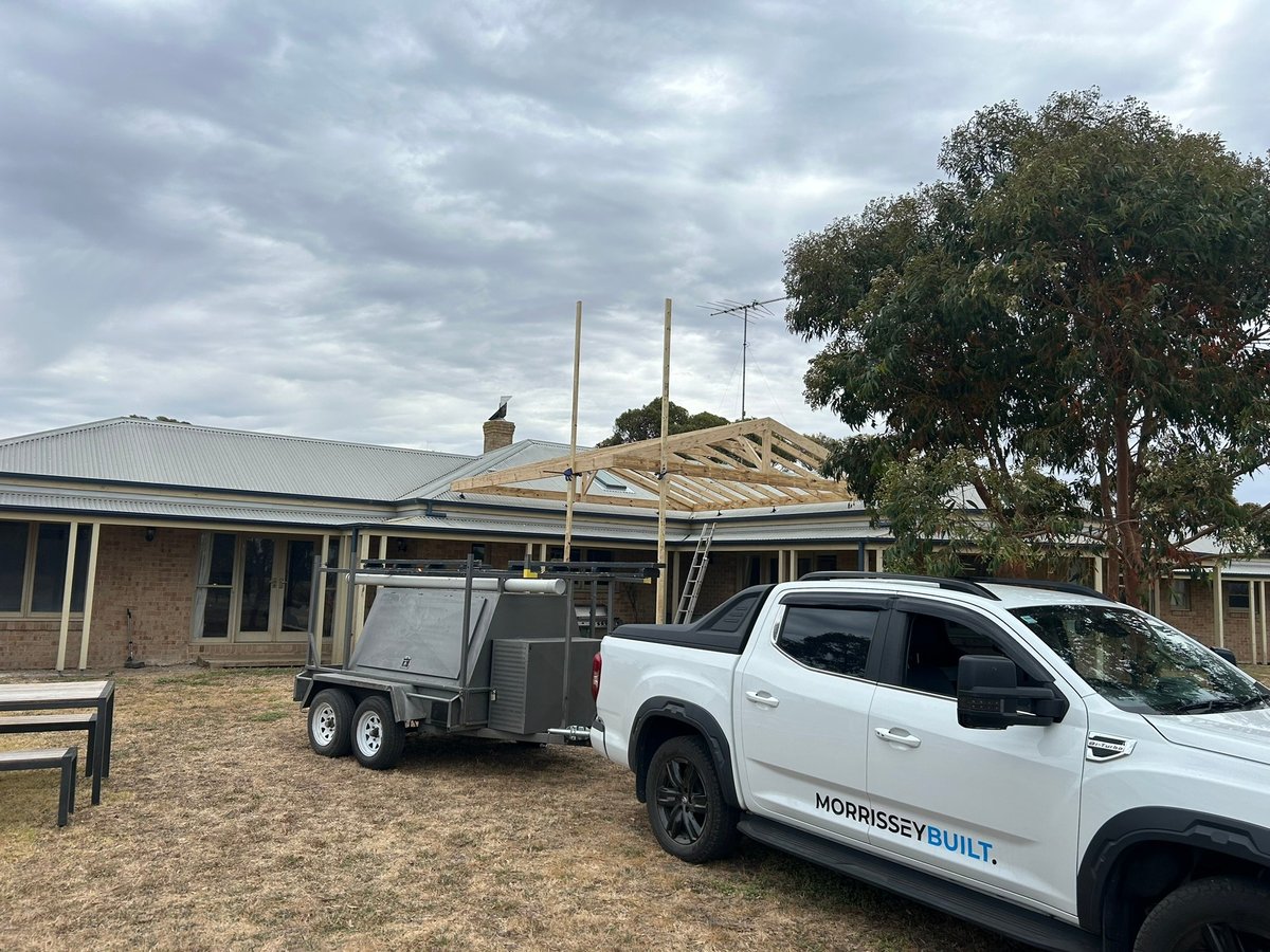 White ute with roof frame construction loaded on trailer next to residential house under cloudy sky