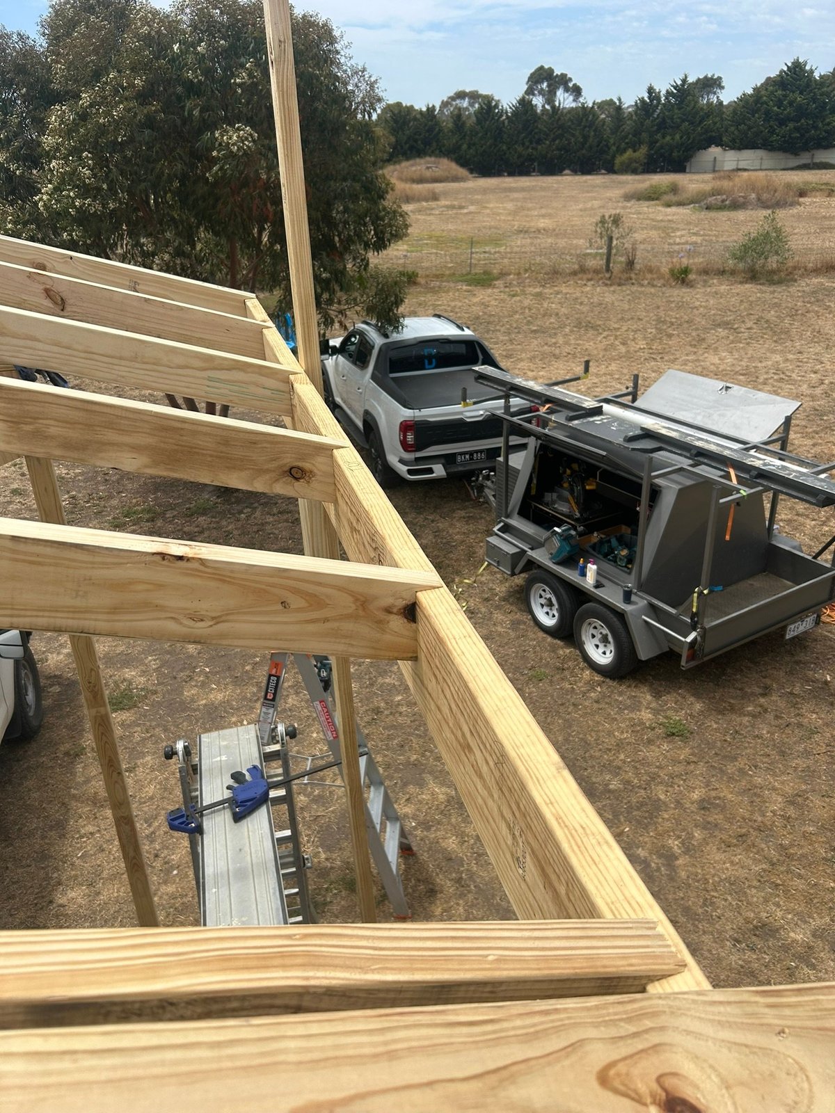 Wooden frame structure under construction with truck and enclosed trailer in rural field setting