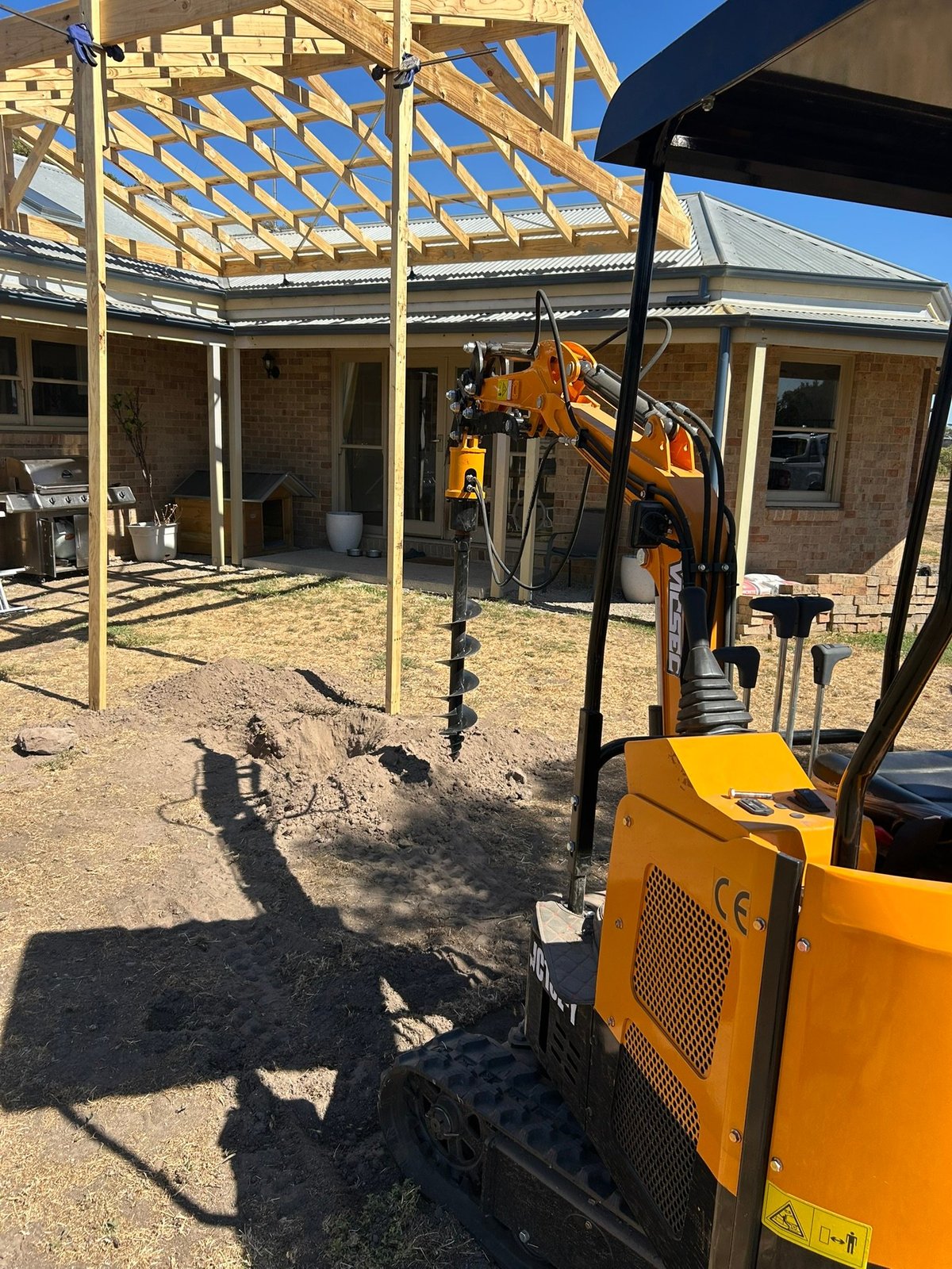 Construction site with yellow excavator under timber pergola structure attached to brick building