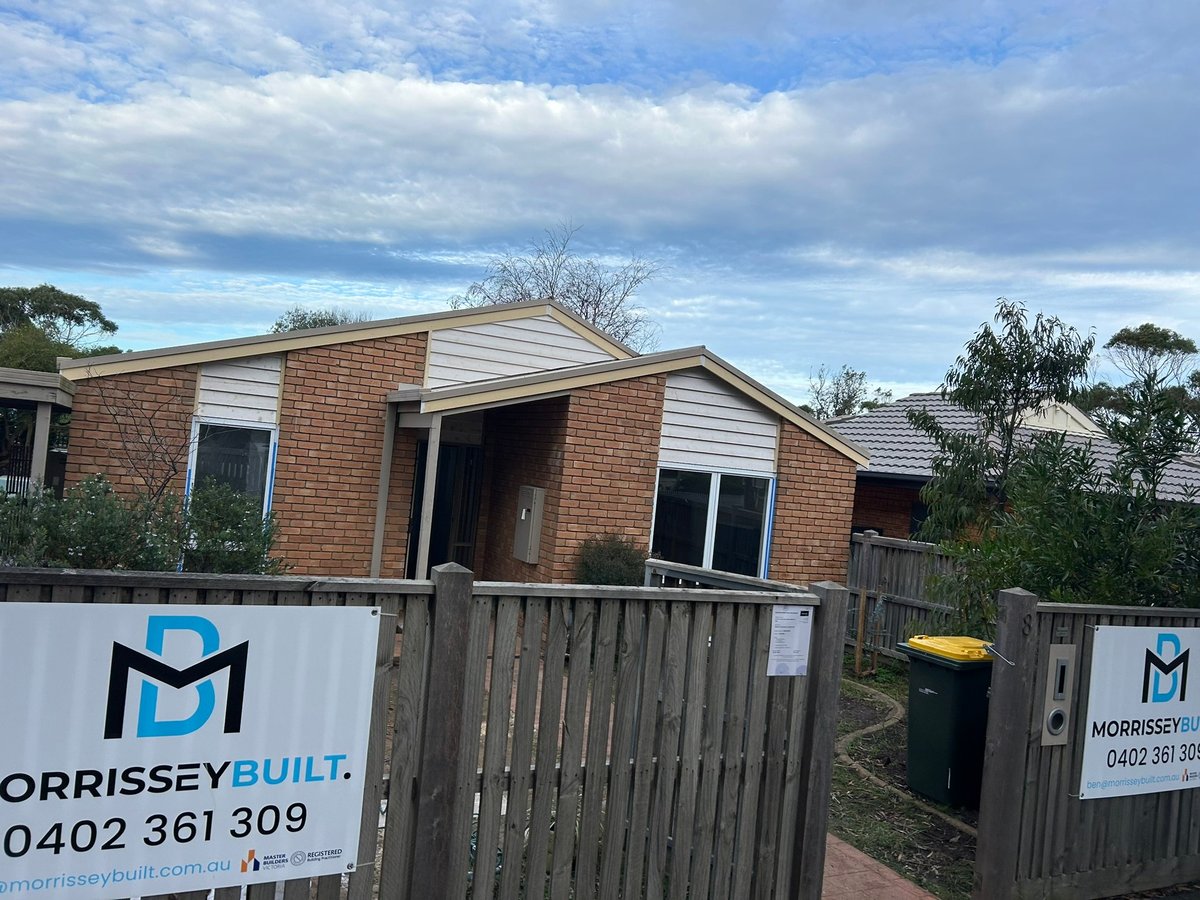 Modern brick residential home with wooden deck and white fence, featuring Morrissey Built signage and blue sky with clouds