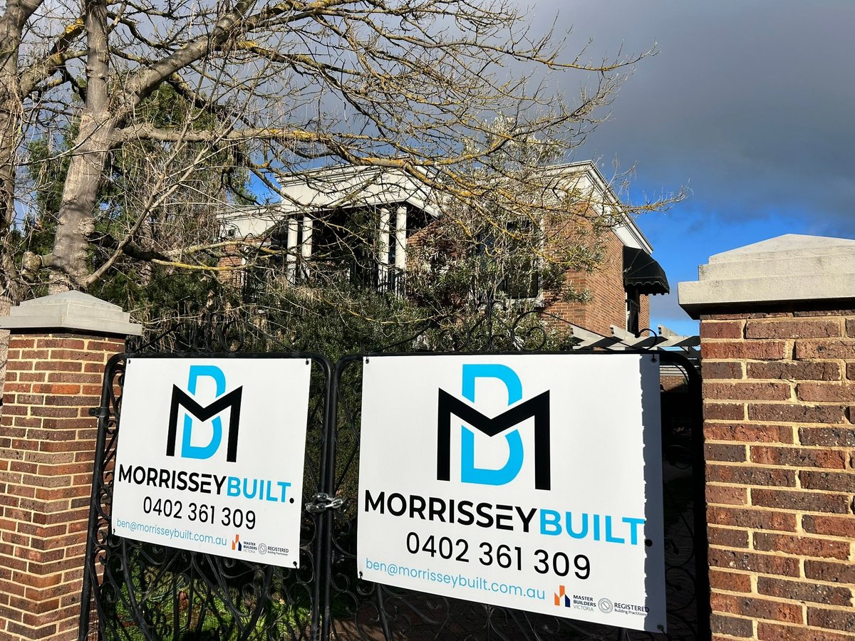 Two Morrissey Built construction company signs attached to a brick gate, with a residential property and bare tree visible in the background