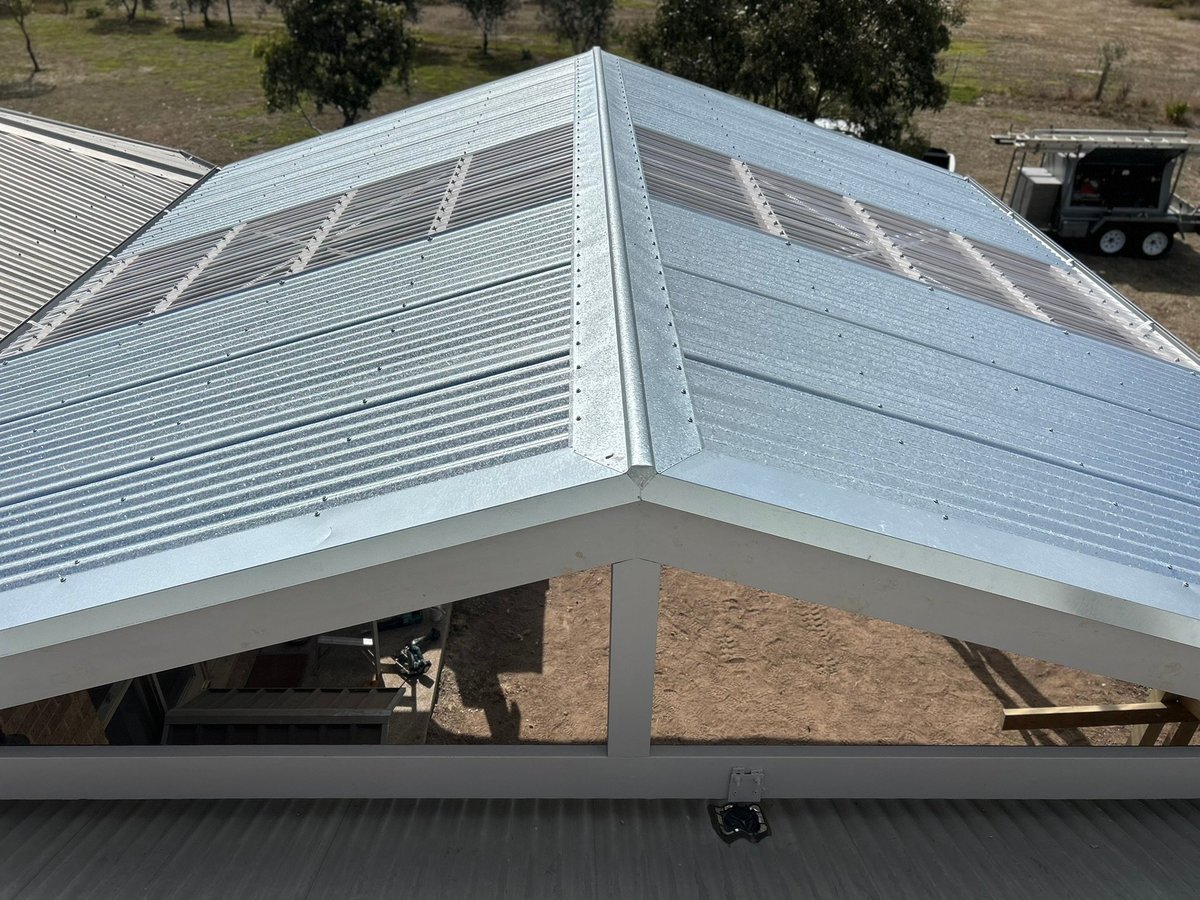 White metal pergola with translucent louvered roof panels attached to a building, viewed from below with trees and equipment visible in background